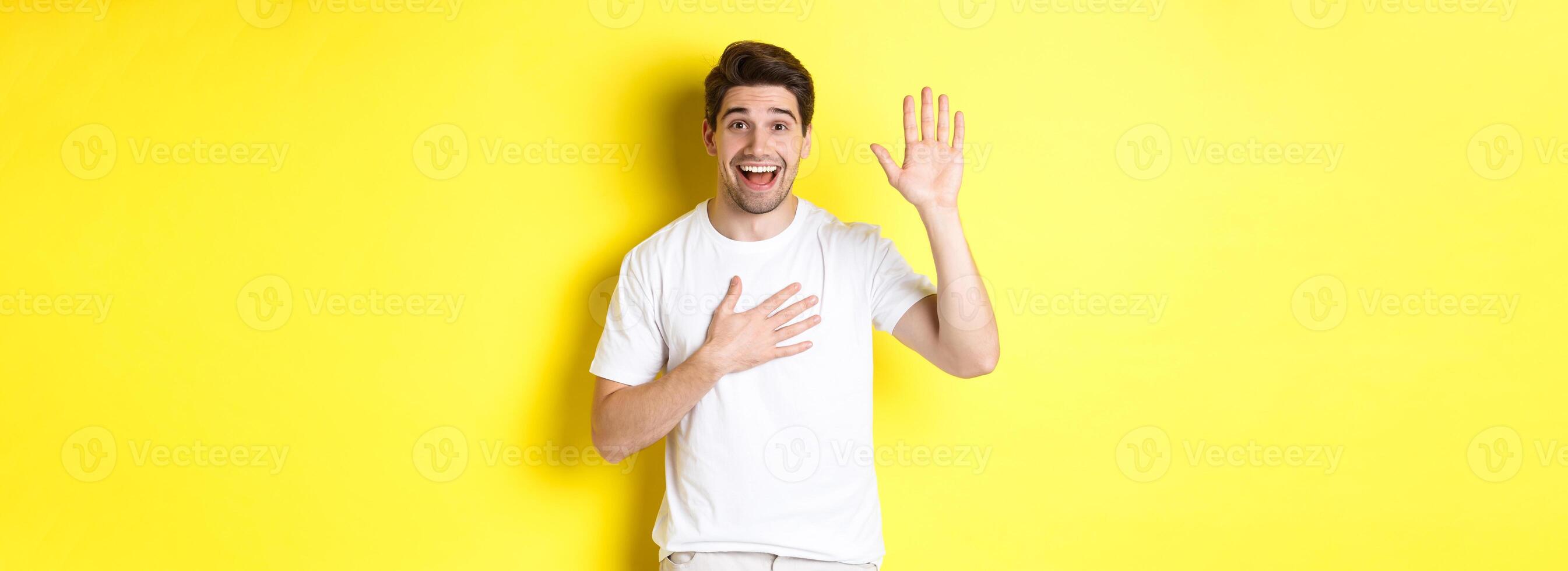 Happy guy making promise, holding hand on heart, swearing to tell truth, standing over yellow background in white t-shirt photo