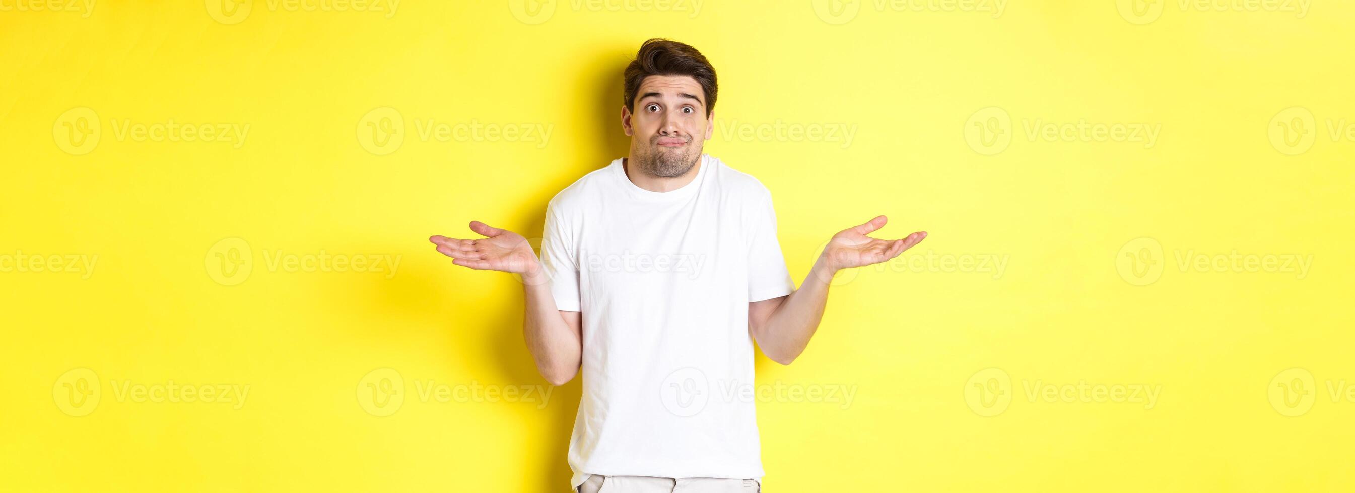 Clueless man shrugging, spread hands sideways confused, dont know anything, standing over yellow background photo