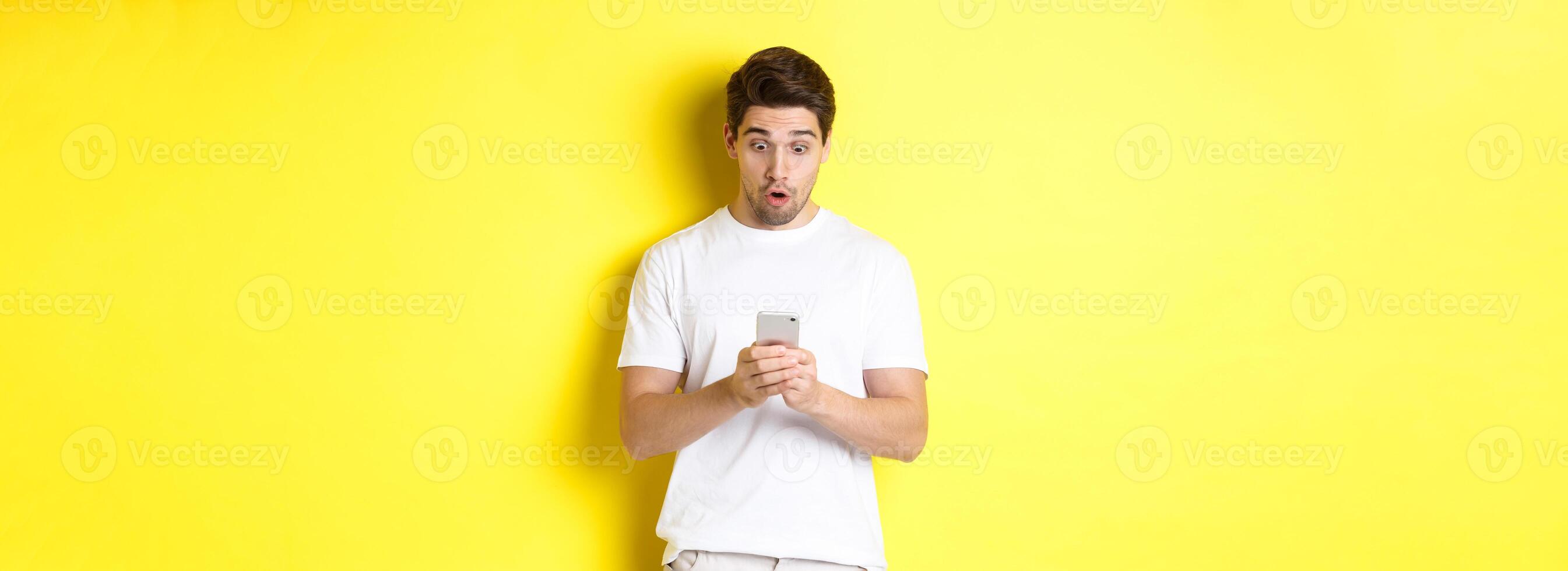 Man looking surprised in smartphone, reading message on cell phone, standing in white outfit against yellow background photo