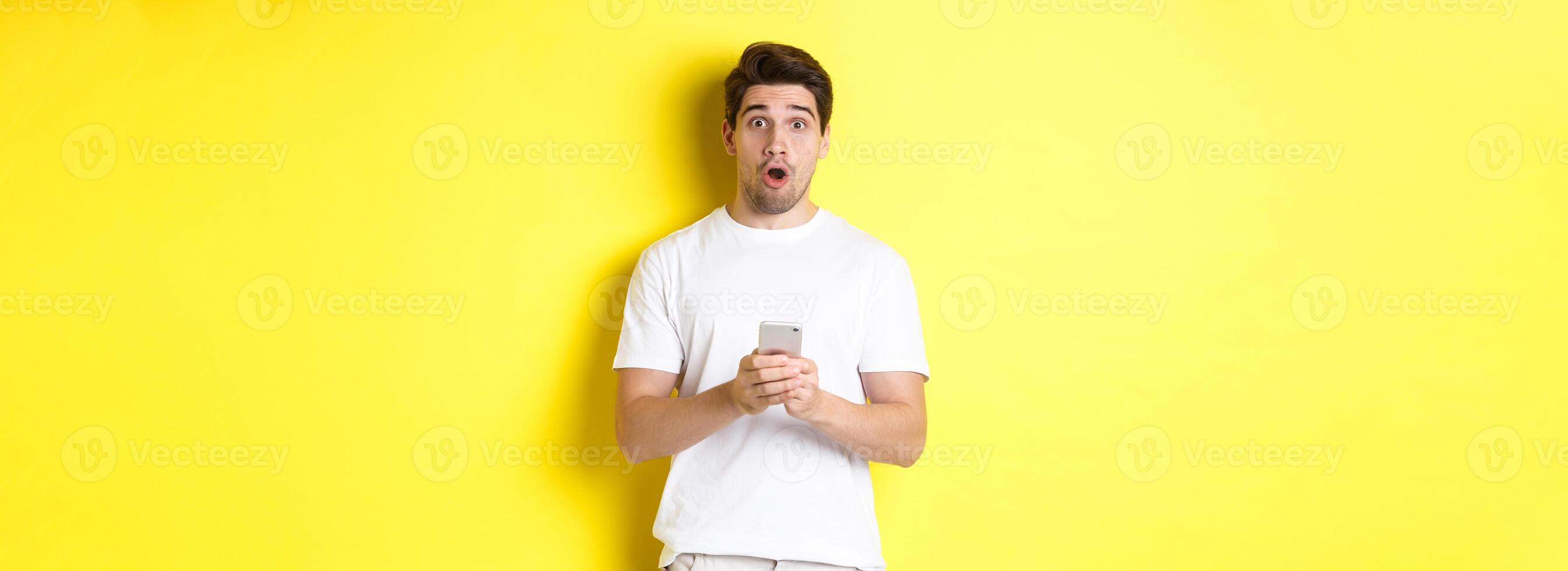Man looking surprised, using smartphone, open mouth and saying wow, standing against yellow background photo