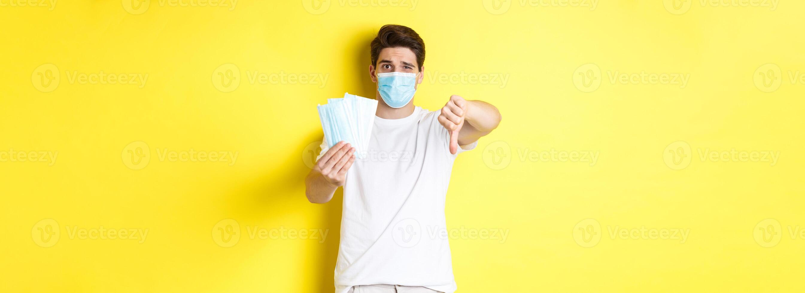 Concept of covid-19, quarantine and preventive measures. Man looking disappointed and showing thumb down, do not recommend bad medical masks, standing over yellow background photo