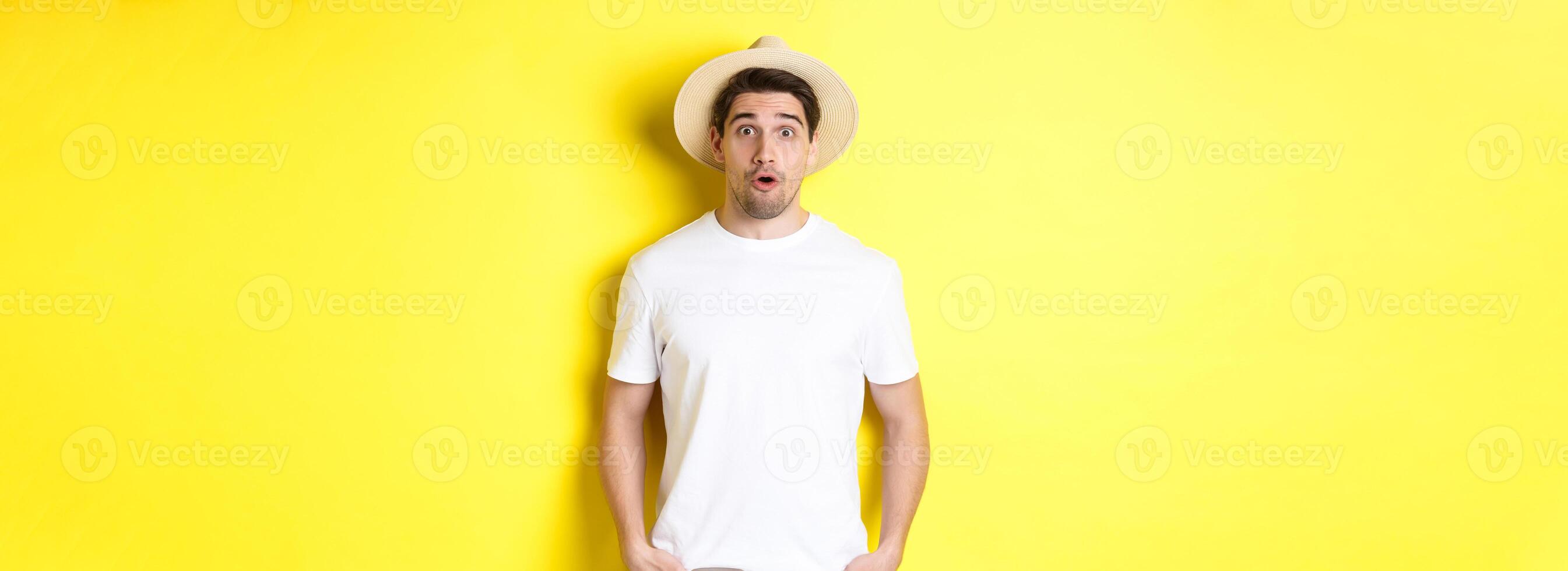 Concept of travelling and tourism. Surprised caucasian guy in straw hat, say wow, standing against yellow background photo