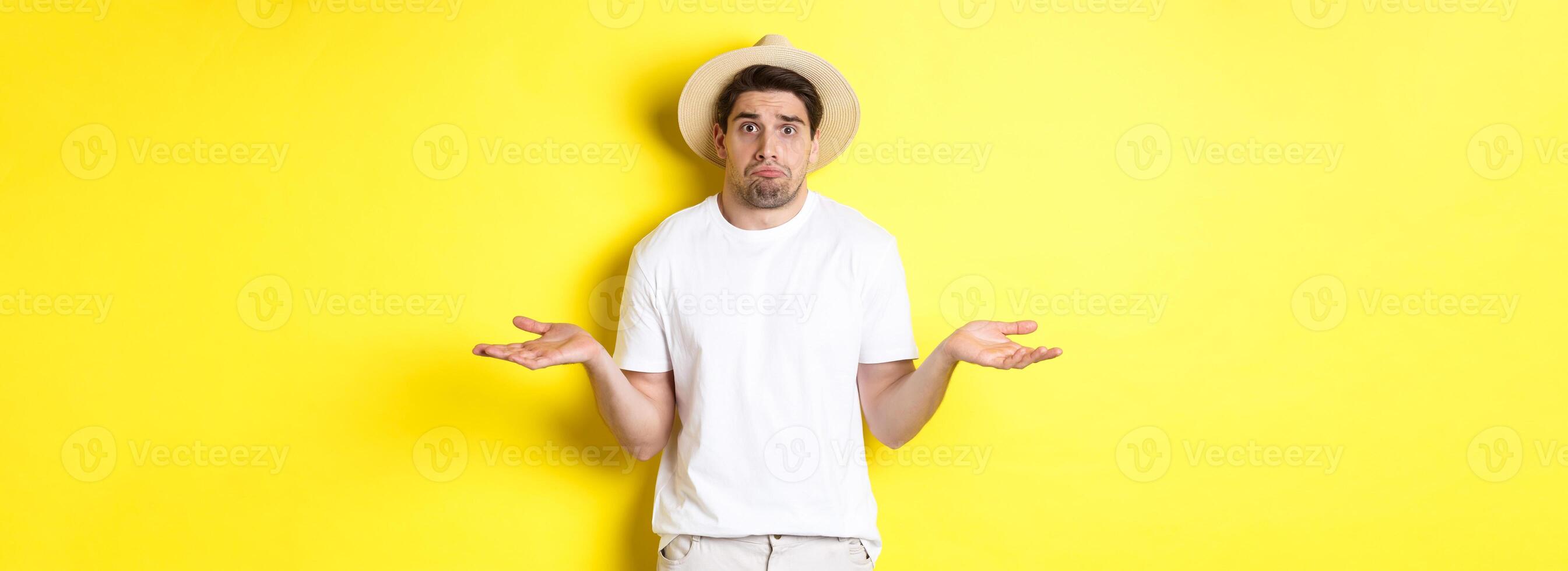 Concept of tourism and summer. Confused male tourist shrugging, looking indecisive, standing against yellow background photo