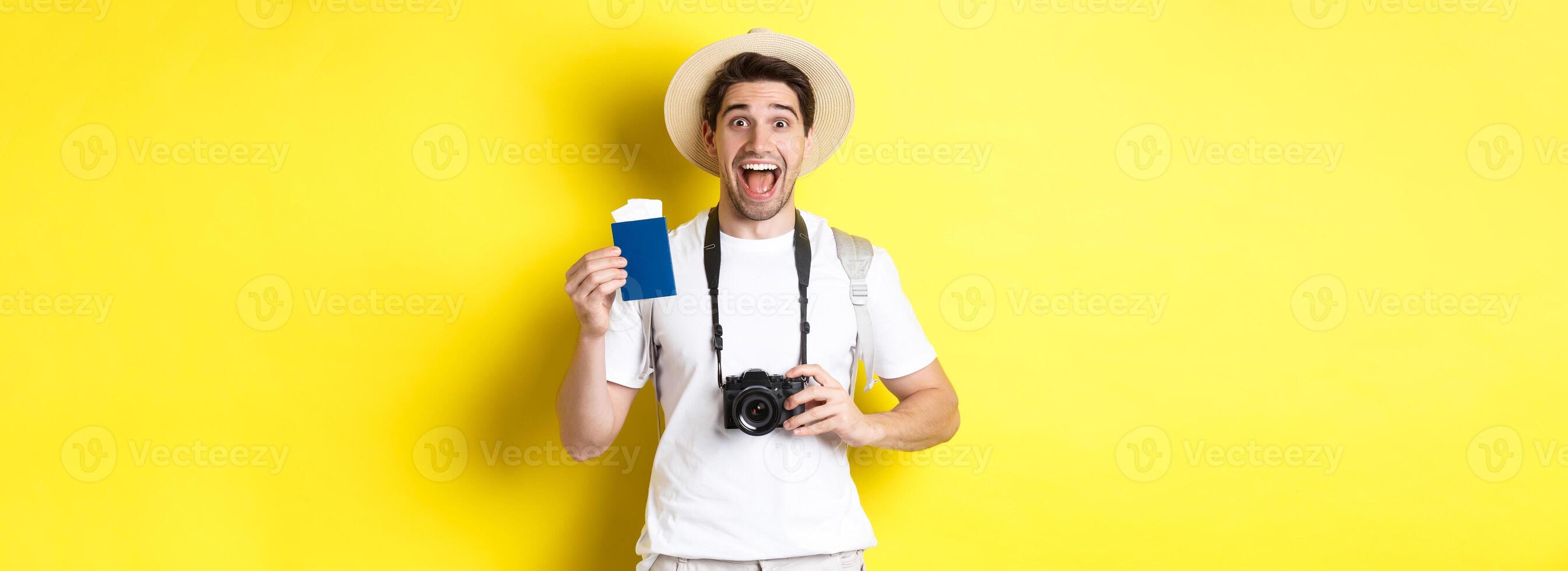 Travelling, vacation and tourism concept. Excoted tpirost showing passport with tickets, holding camera and wearing straw hat, standing over yellow background photo