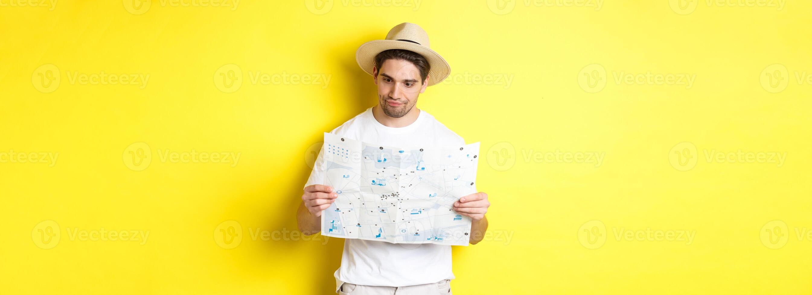 Travelling, vacation and tourism concept. Male tourist looking confused at map, looking for right direction, standing over yellow background photo