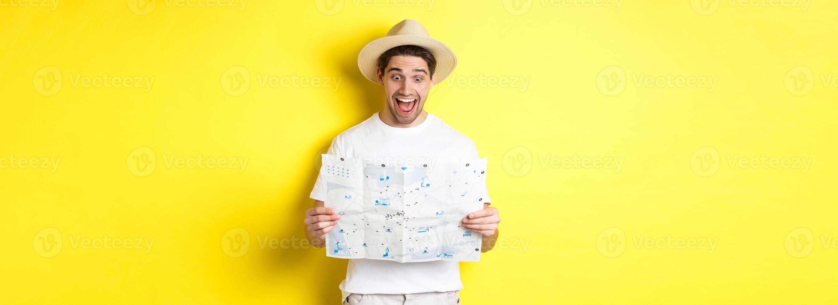Travelling, vacation and tourism concept. Man tourist looking happy at map with sighsteeings, explore city, standing over yellow background photo