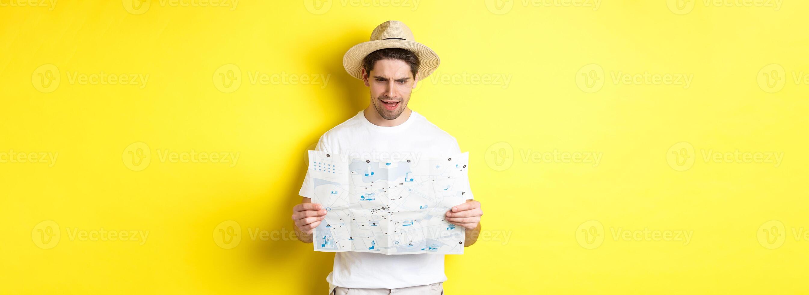 Travelling, vacation and tourism concept. Man looking confused at map during trip, cant understand, standing over yellow background photo