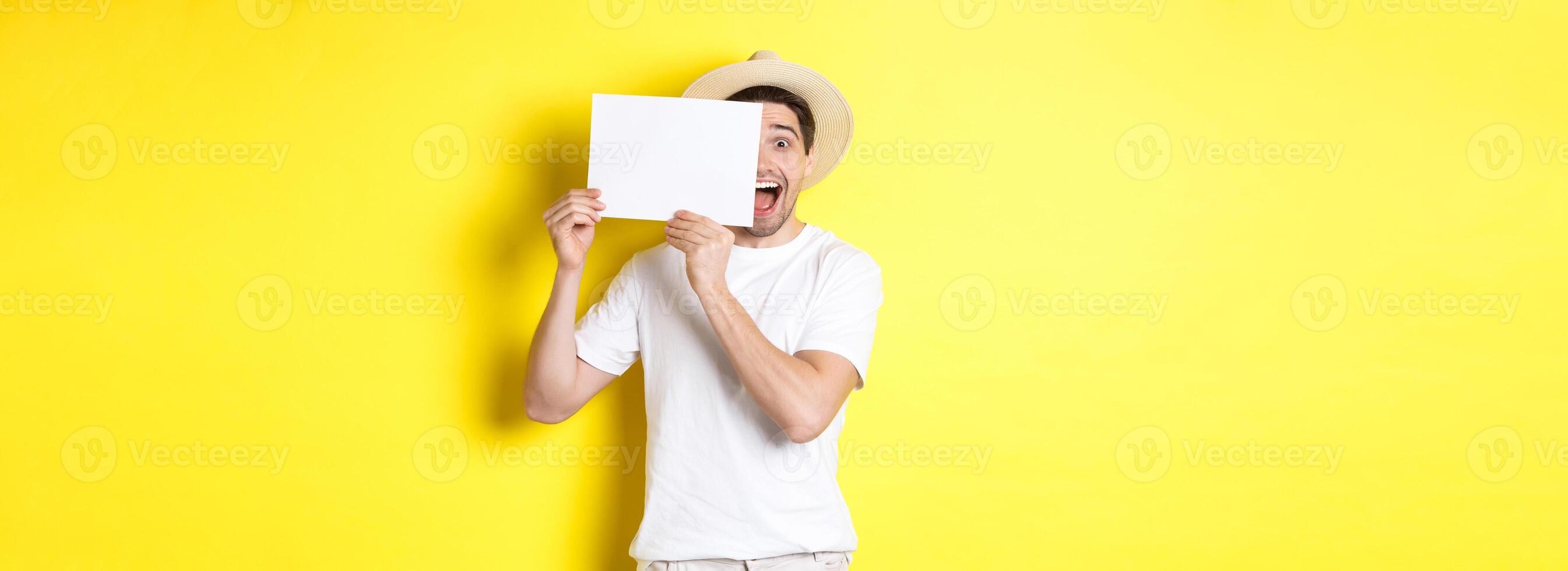 Excited tourist on vacation showing blank piece of paper for your logo, holding sign near face and smiling, standing against yellow background photo