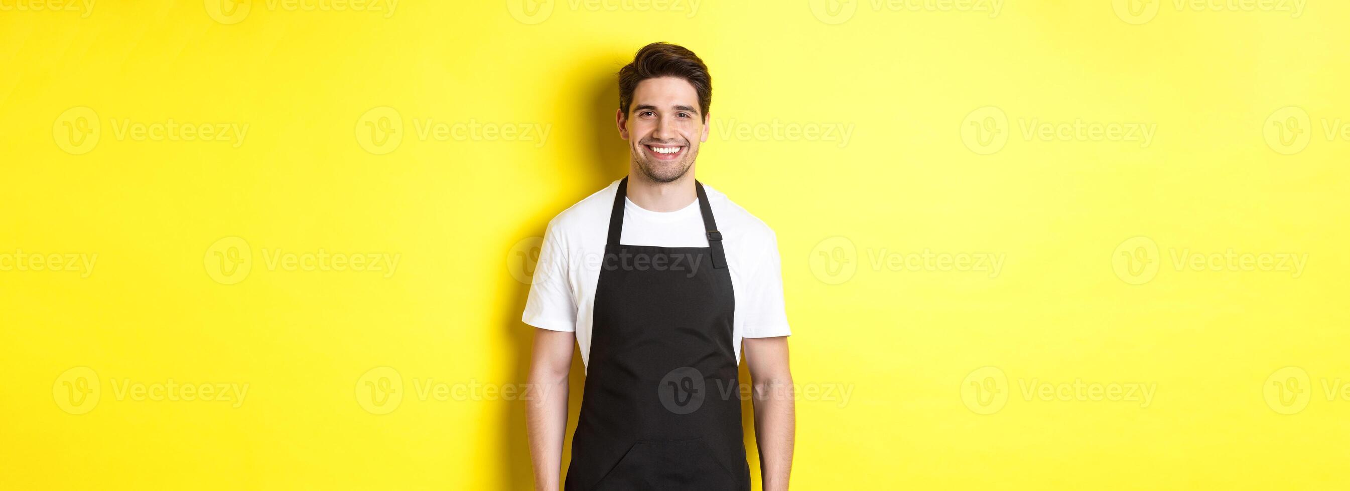 Young man waiter in black apron, smiling, working in store or coffee shop, standing against yellow background photo