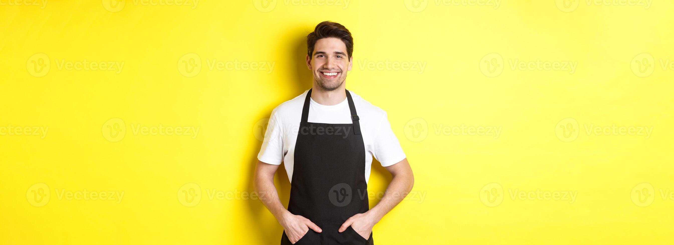Happy barista in black apron looking at camera. Coffee shop owner wearing cafe uniform and smiling, standing over yellow background photo
