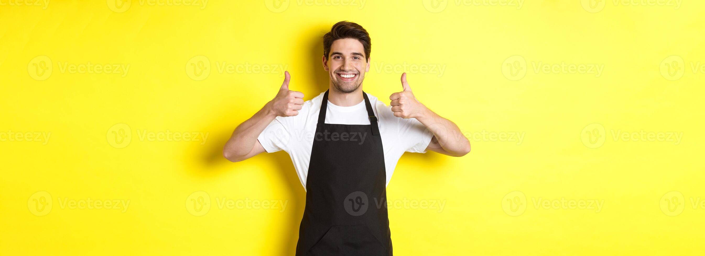 Smiling seller in black apron showing thumbs up, approve or like something, recommending cafe or store, yellow background photo