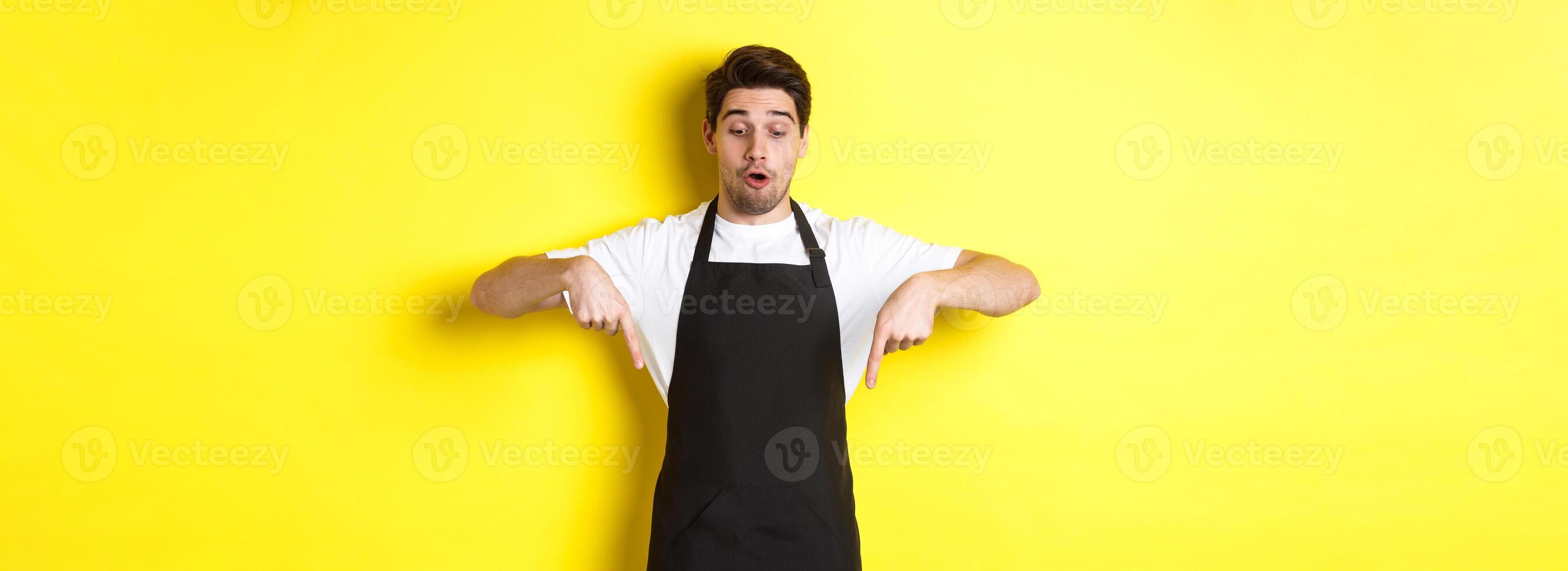 Surprised male barista in black apron checking out promo offer, pointing fingers down at logo banner, standing over yellow background photo