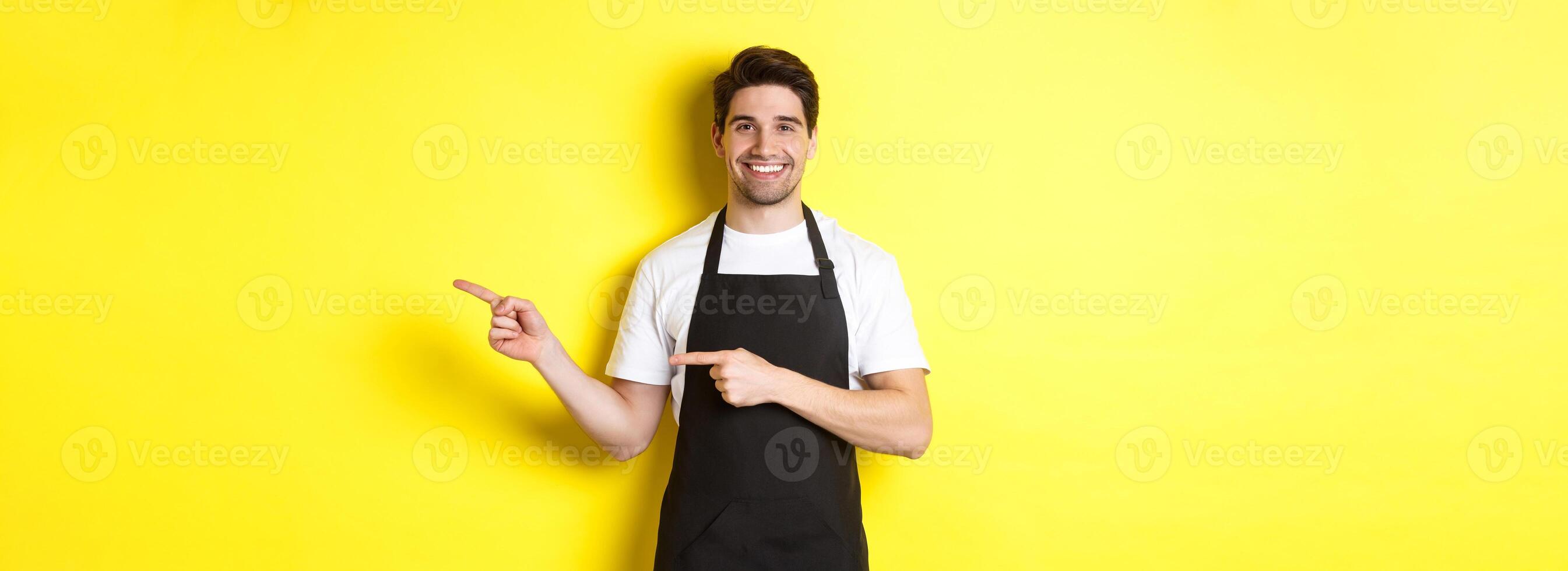 Friendly waiter pointing fingers right, showing your logo or promo offer, wearing black apron uniform, standing over yellow background photo