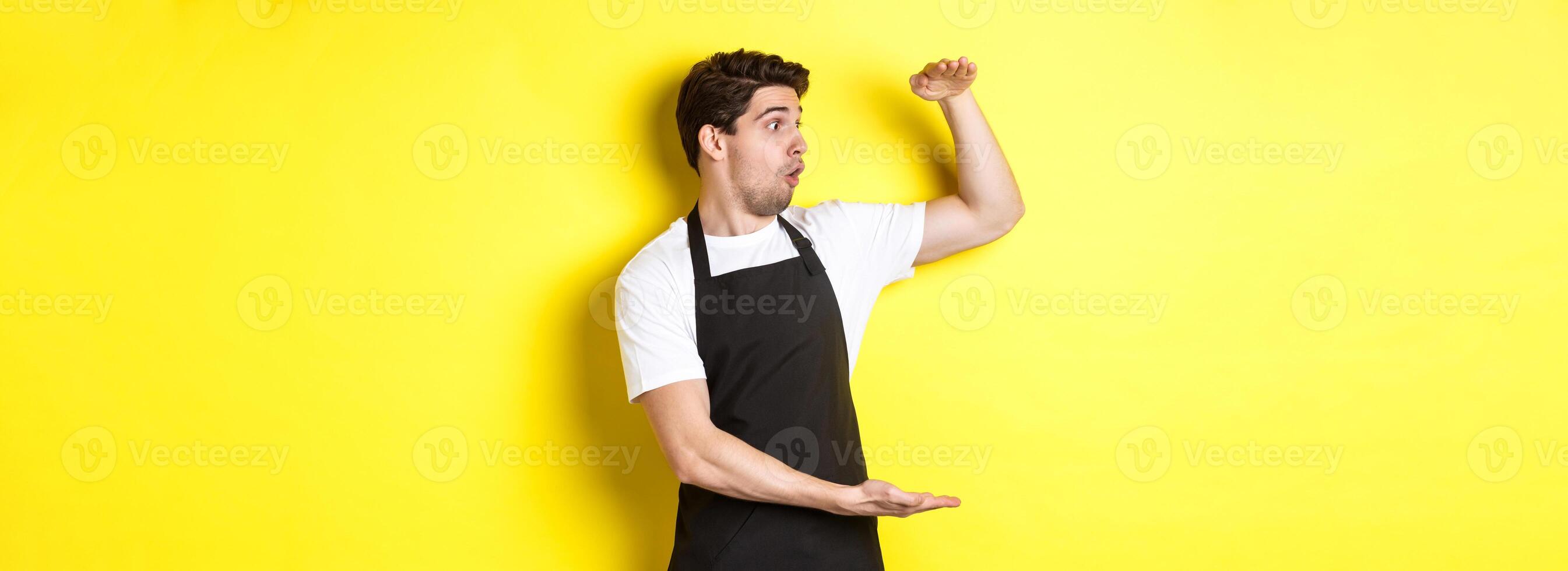 Waiter in black apron looking amazed at something large, holding big object, standing over yellow background photo