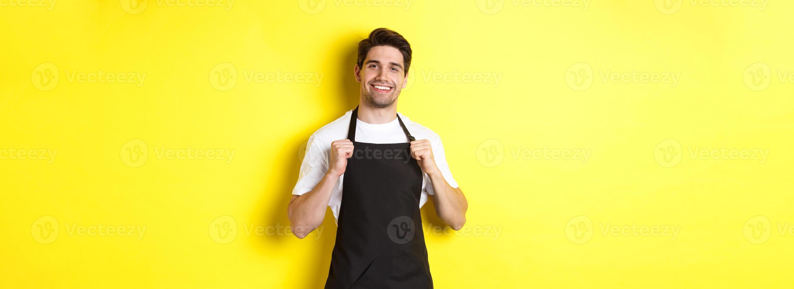 Confident barista in black apron standing against yellow background. Waiter smiling and looking happy photo