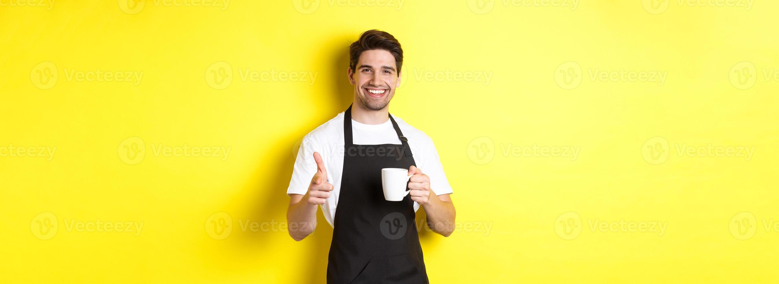 Barista bringing coffee and pointing finger gun at camera, standing in black apron against yellow background photo