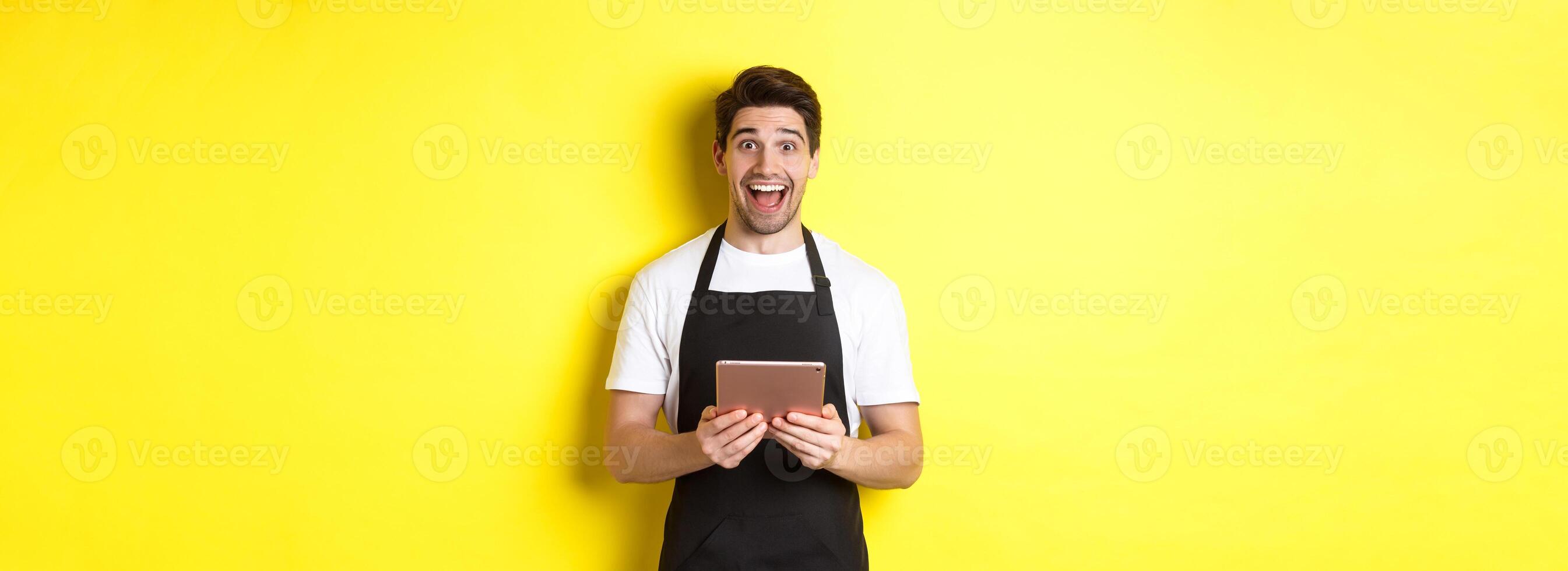 Happy salesman in black apron, holding digital tablet and looking surprised, standing against yellow background photo