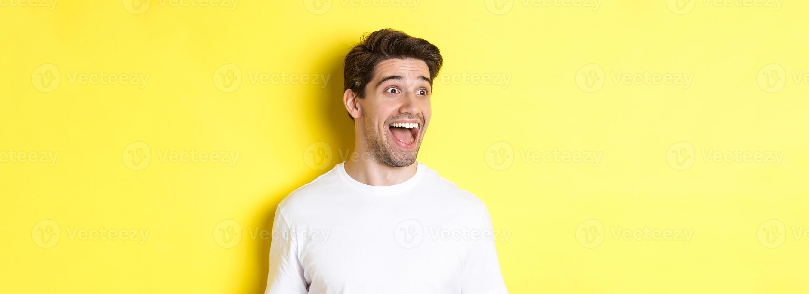 Image of happy man checking out promo, looking left with amazement, standing in white t-shirt against yellow background photo