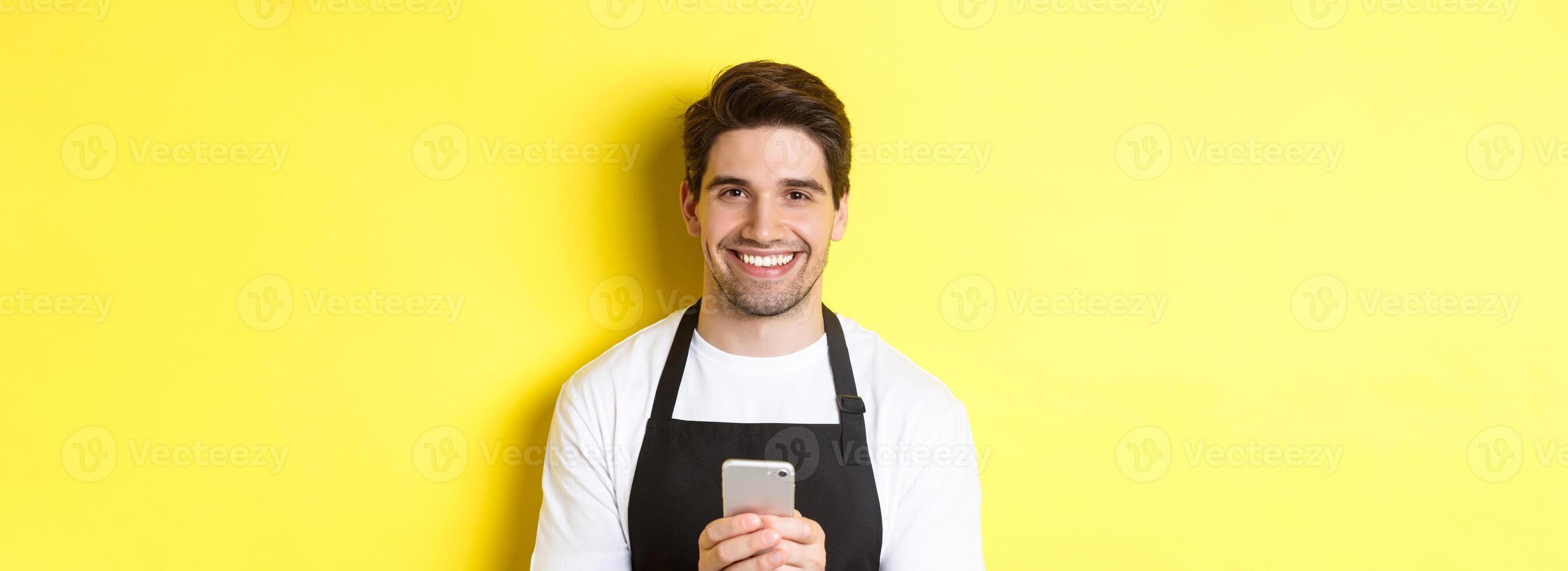 Close-up of handsome barista sending message on mobile phone, smiling happy, standing over yellow background photo