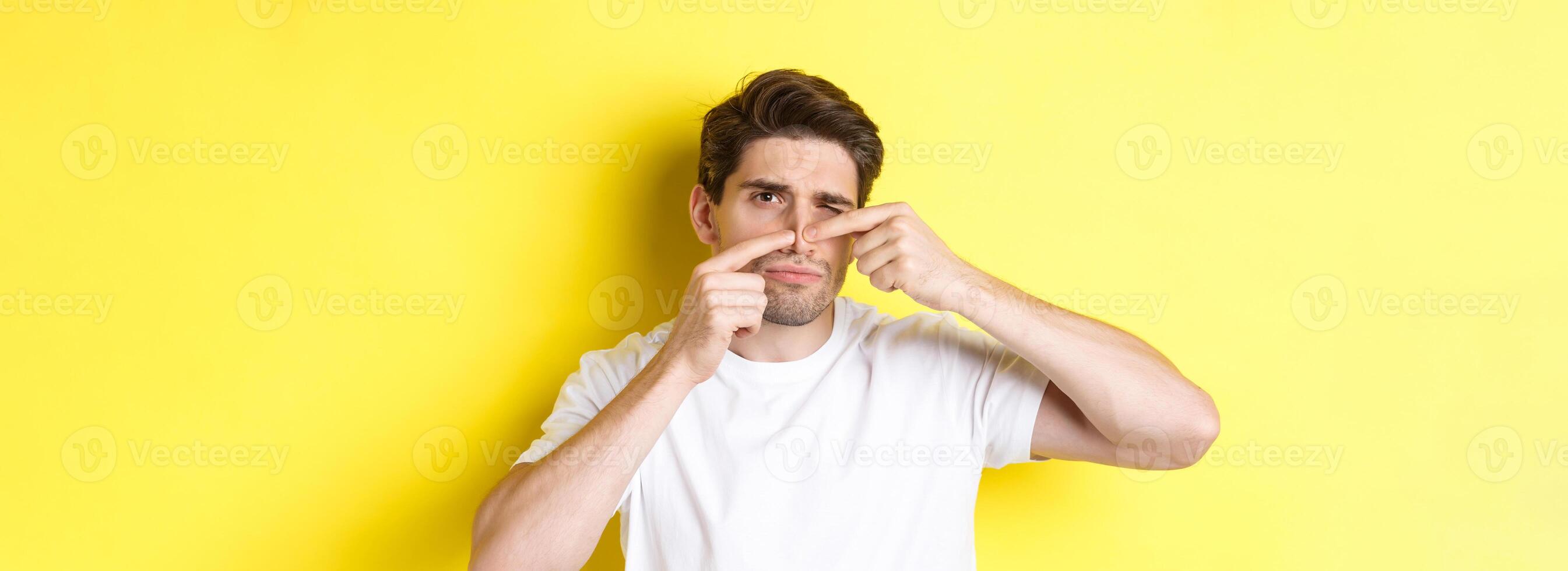 Young man squeezing pimple on nose, standing over yellow background. Concept of skin care and acne photo