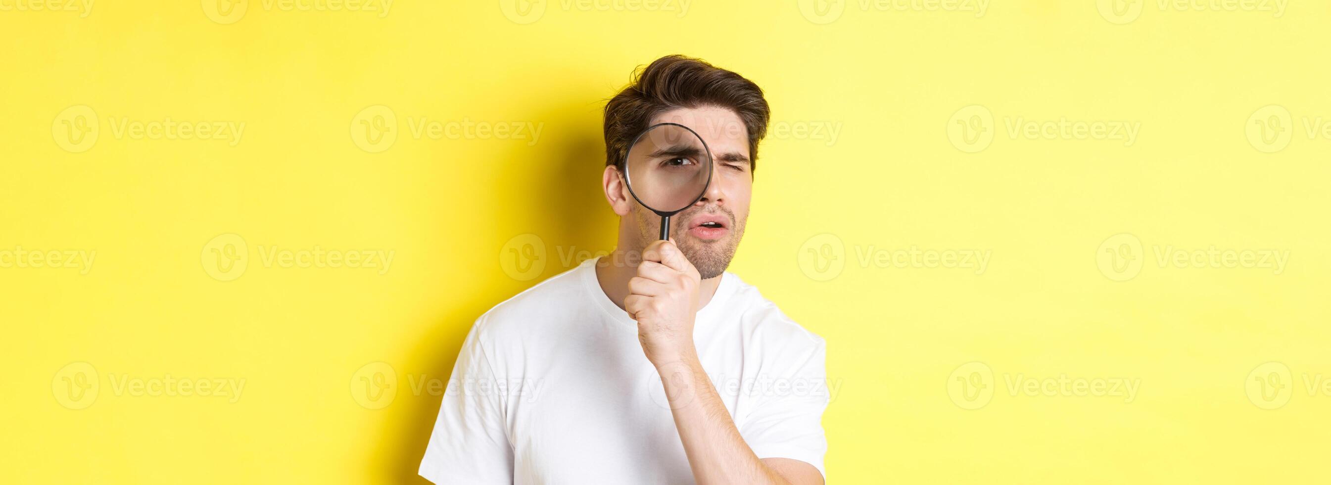 Man looking through magnifying glass with serious and thoughtful look, searching or investigating, standing over yellow background photo