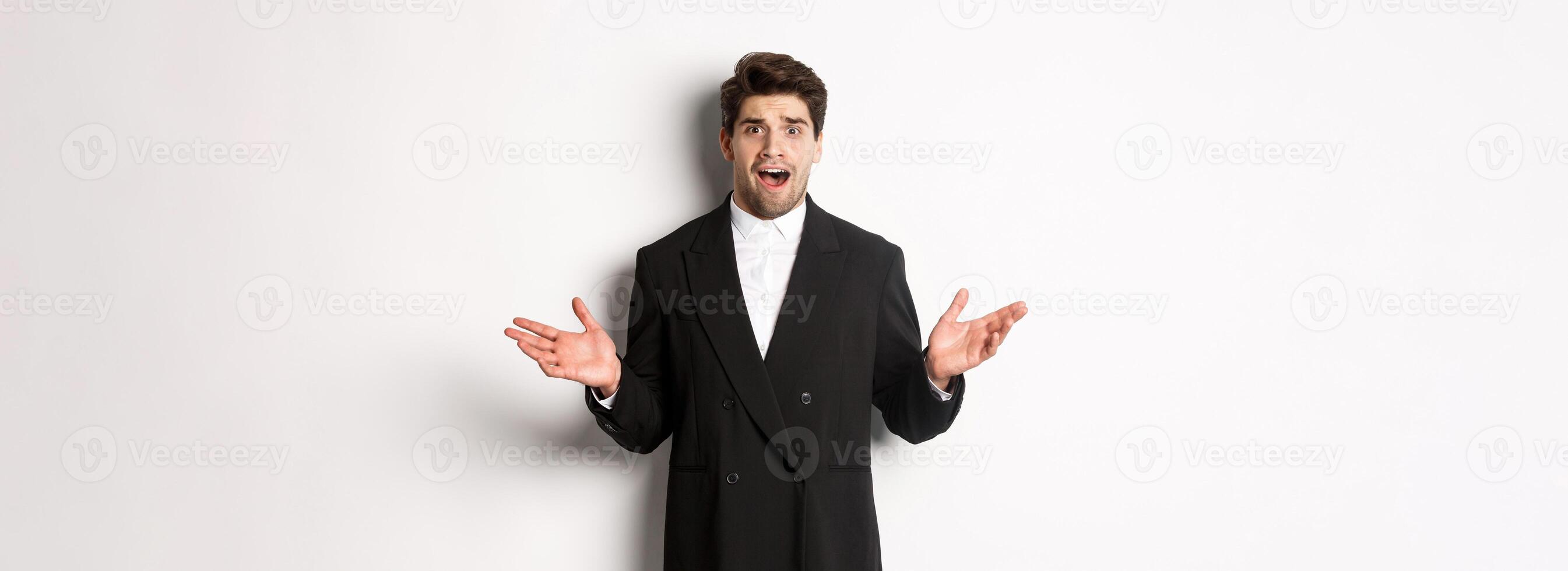 Portrait of confused and worried handsome man in suit, looking at something strange, spread hands sideways and standing puzzled against white background photo