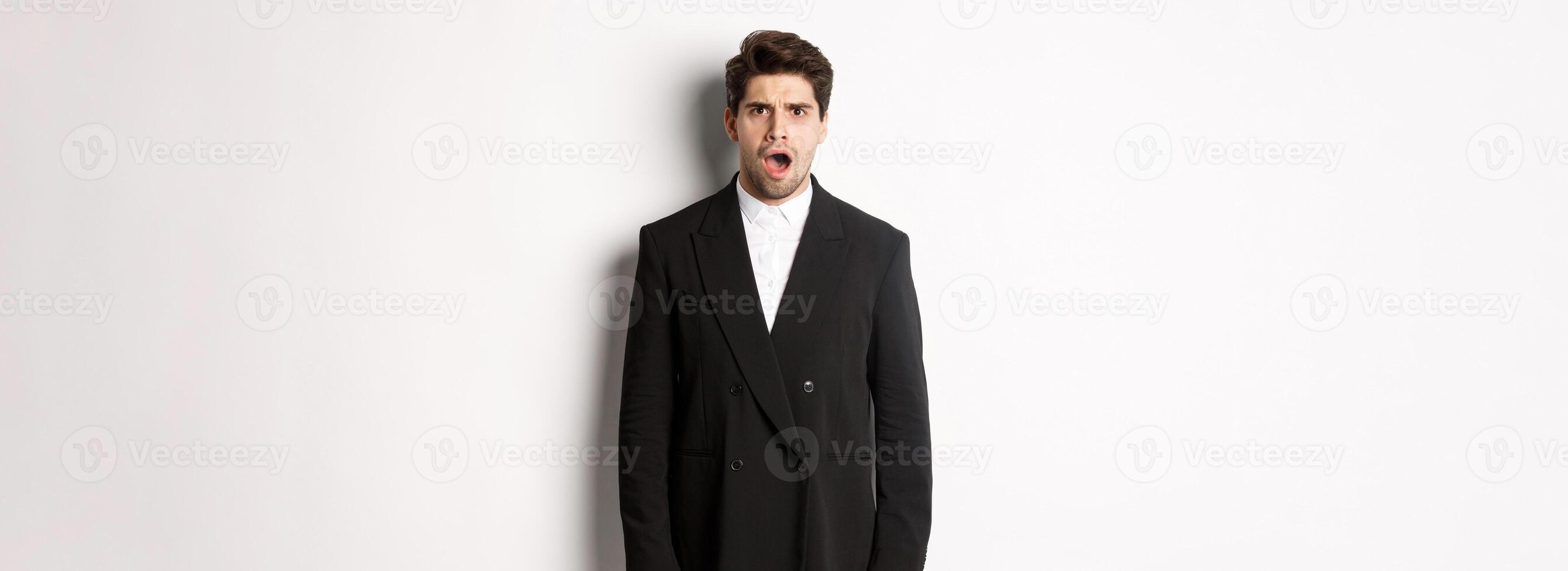 Portrait of shocked and startled handsome man in suit, drop jaw and looking in awe at camera, standing over white background photo