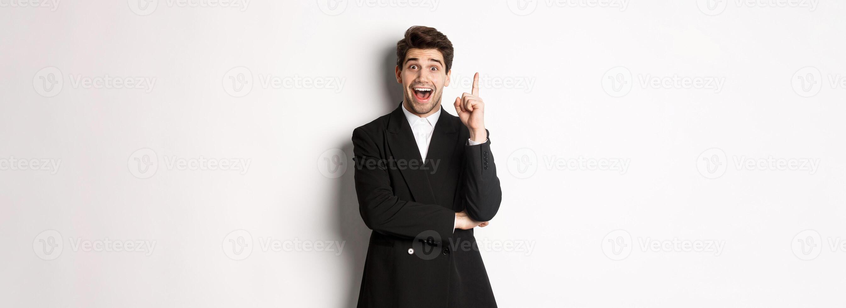 Portrait of attractive man in suit, having an idea, standing excited and raising one finger to tell suggestion, think-up solution, standing over white background photo