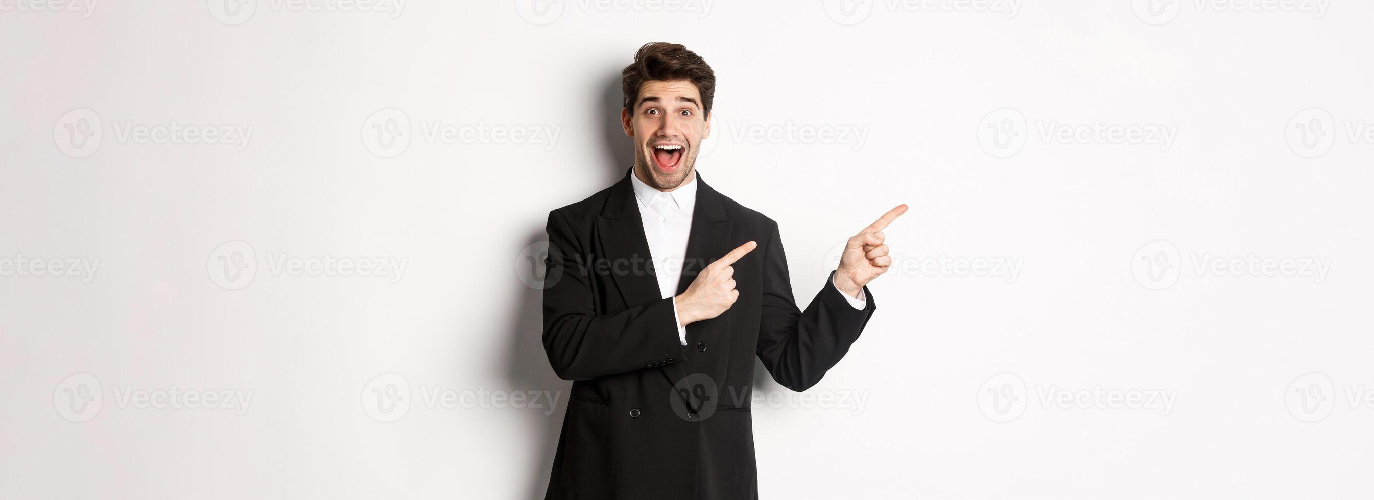 Portrait of excited and happy man in suit showing advertisement, pointing fingers at upper left corner and smiling amazed, standing over white background photo