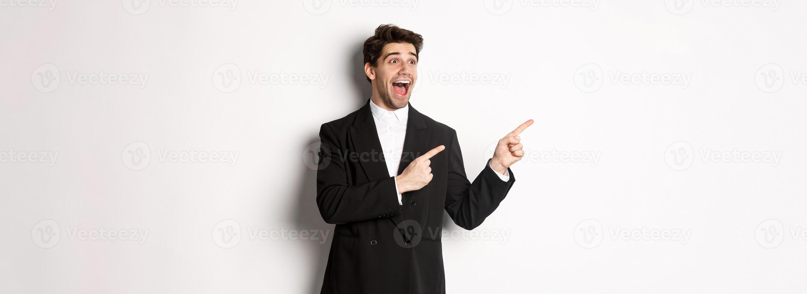 Portrait of amazed and happy handsome man in black suit, pointing and looking left impressed, showing awesome christmas promo, standing over white background photo
