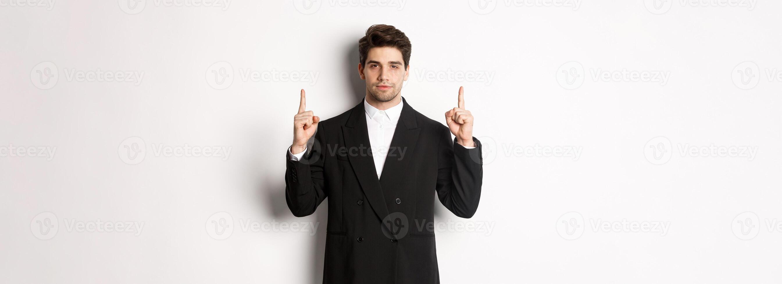 Image of confident and handsome man in formal suit, pointing fingers up, showing copy space on white background photo