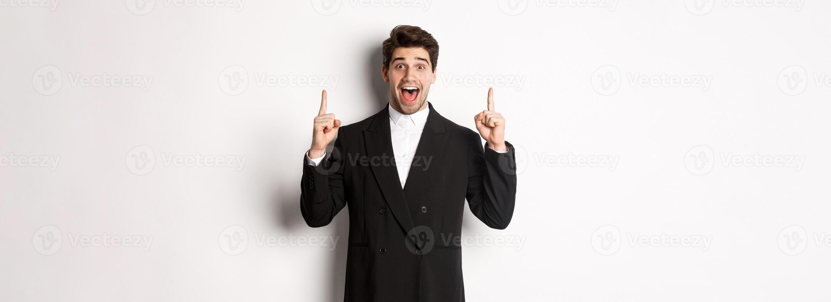 Image of handsome man in party suit, showing holidays promo, pointing fingers up and smiling amazed, standing over white background photo