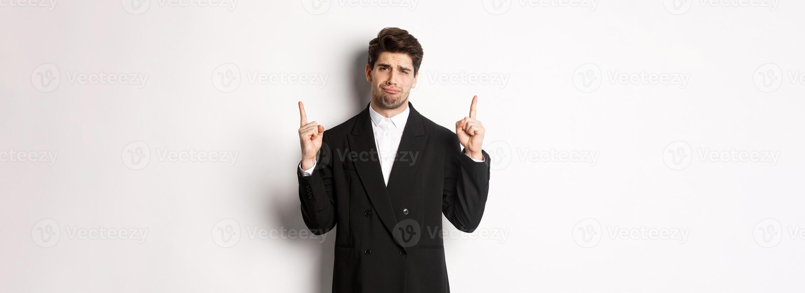 Portrait of reluctant and sad handsome man, complaining and pointing fingers up at something bad, looking jealous, standing over white background photo