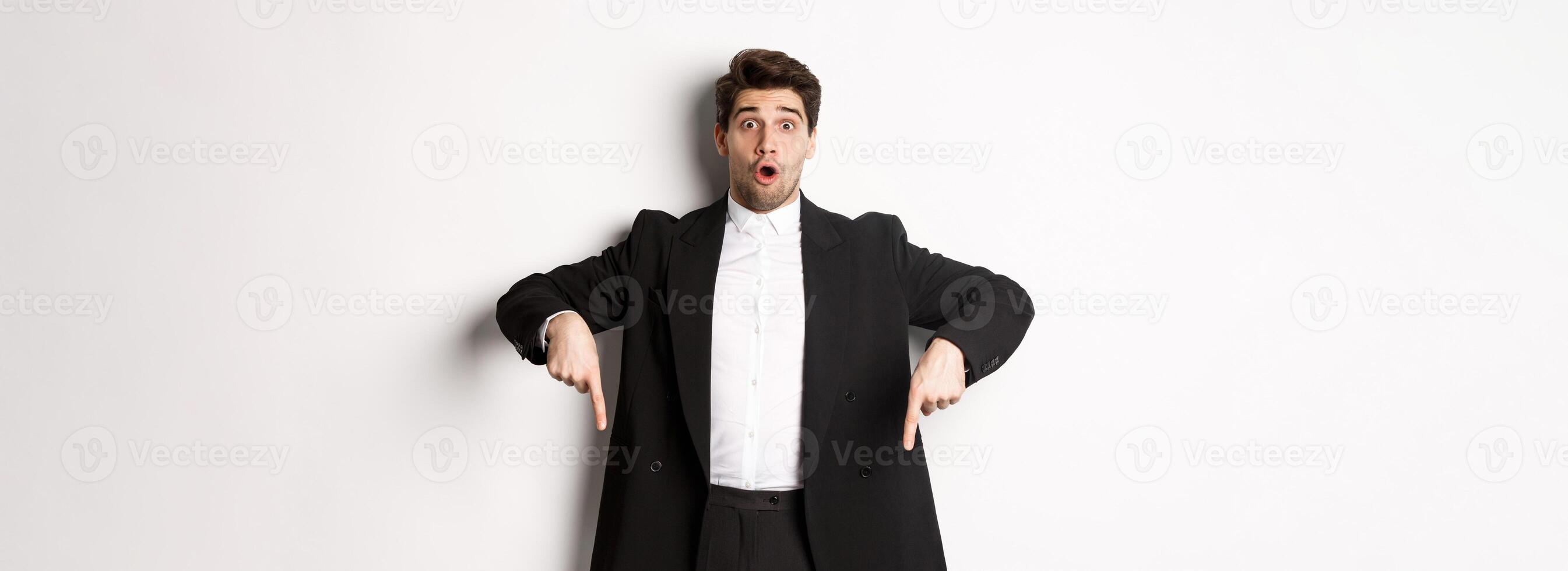 Portrait of surprised and excited man in black suit, pointing fingers down and showing christmas advertisement, standing over white background photo