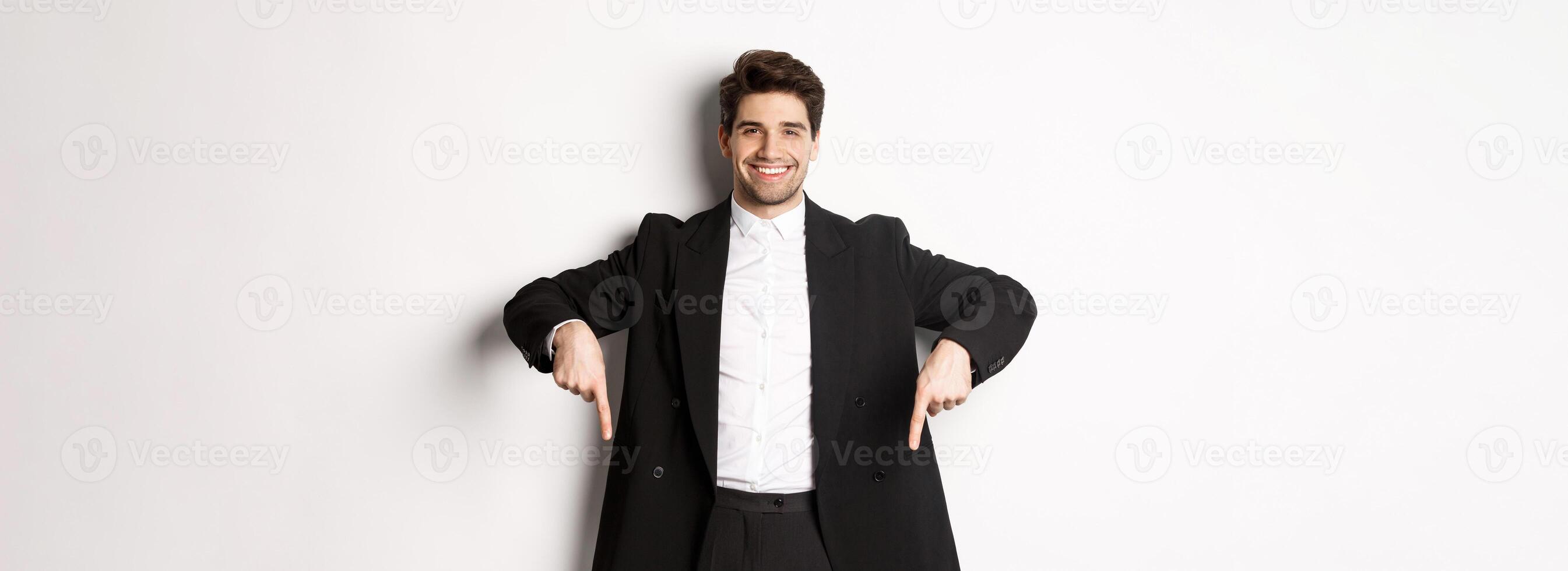 Portrait of good-looking stylish man in black suit, pointing fingers down and smiling, showing winter holidays promo, standing over white background photo