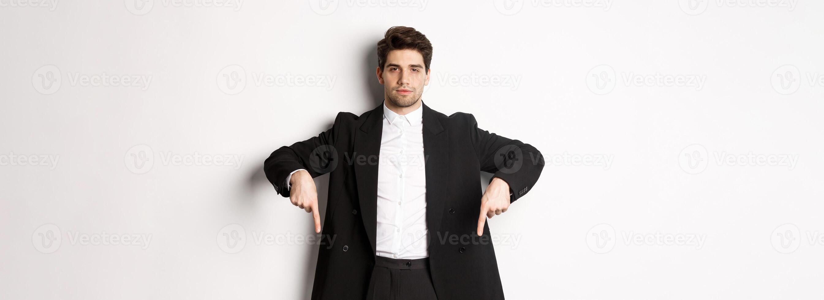 Image of handsome man dressed for formal party, wearing suit and pointing fingers down, showing advertisement or making announcement, standing over white background photo