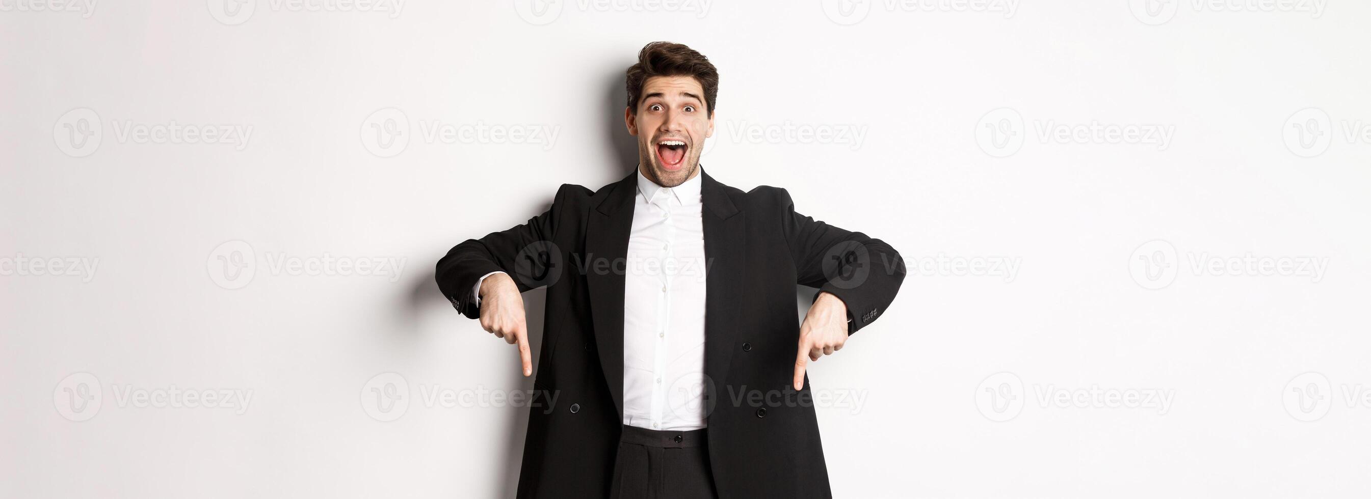 Portrait of super happy man showing christmas promo offer, pointing fingers down and smiling amazed, wearing formal party suit, standing over white background photo