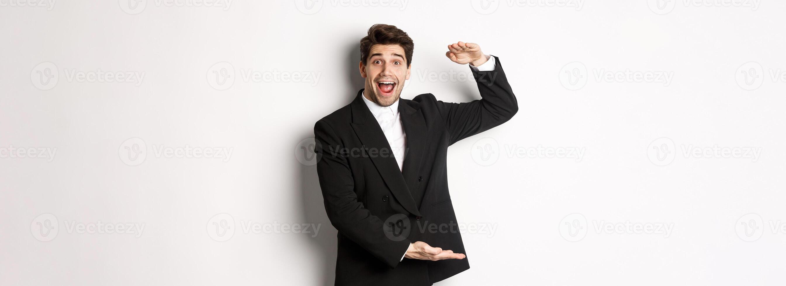 Portrait of excited handsome man in suit, shaping big object on copy space and smiling amazed, holding something, standing over white background photo