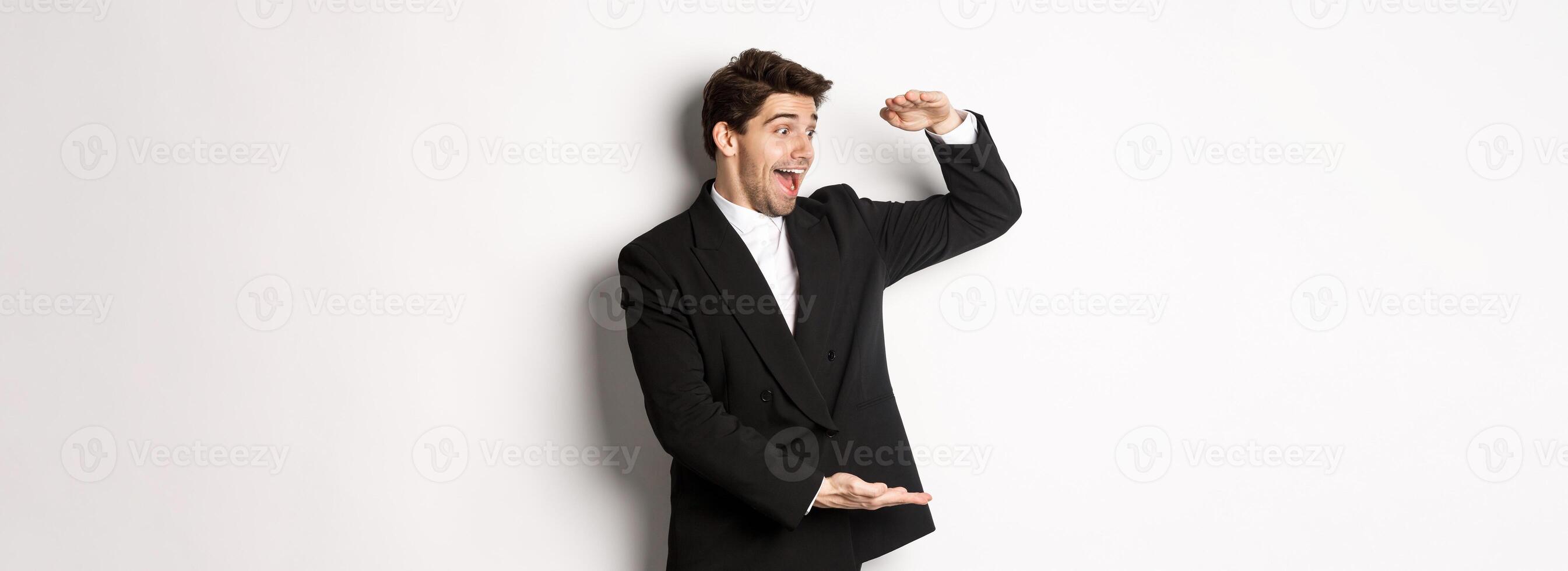 Portrait of excited handsome man in suit, shaping big object on copy space and smiling amazed, holding something, standing over white background photo
