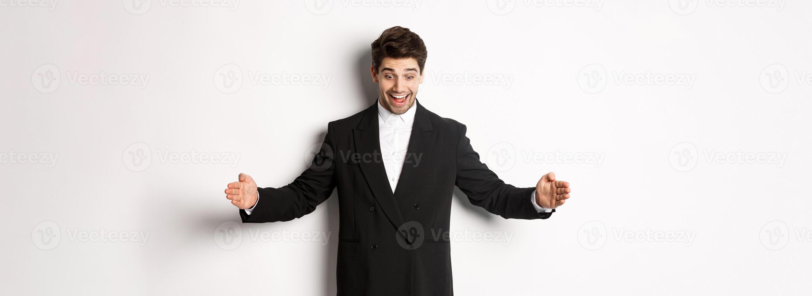 Portrait of excited handsome man in suit, shaping big object on copy space and smiling amazed, holding something, standing over white background photo