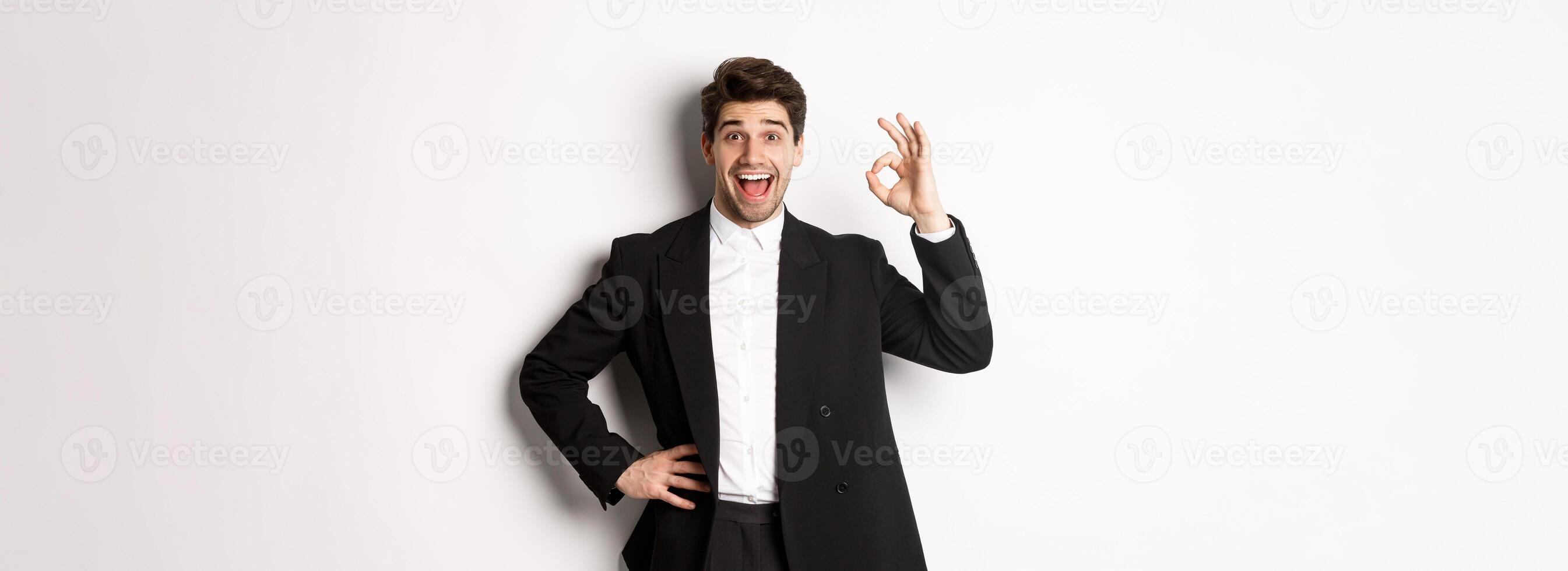 Concept of new year party, celebration and lifestyle. Portrait of pleased handsome guy in black suit, praise something good, showing okay sign in approval, standing over white background photo
