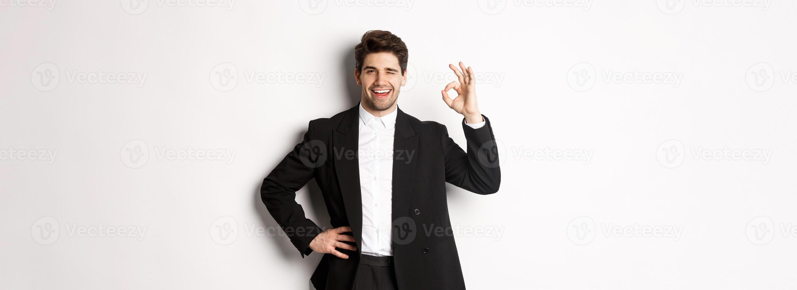 Concept of new year party, celebration and lifestyle. Portrait of confident, successful businessman in suit, showing okay sign and winking, approve something good, white background photo