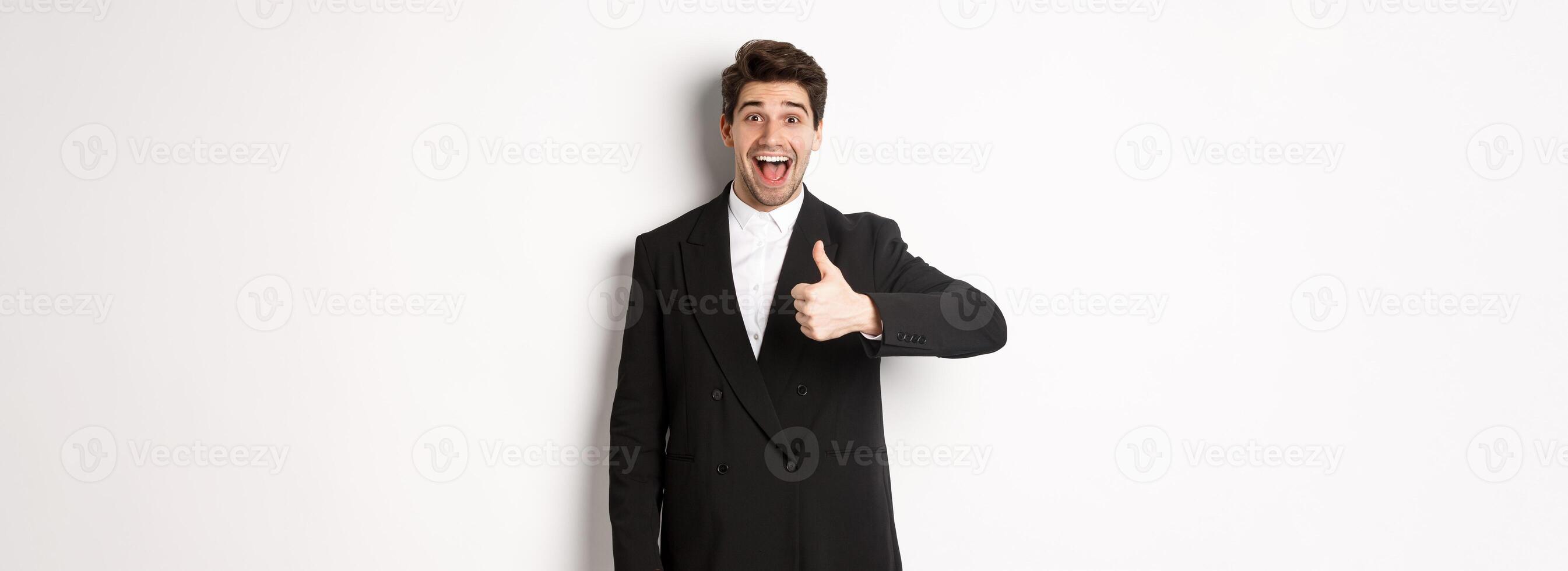 Portrait of amazed and impressed handsome man in black suit, showing thumb-up, praise awesome product, liking something good, standing against white background photo