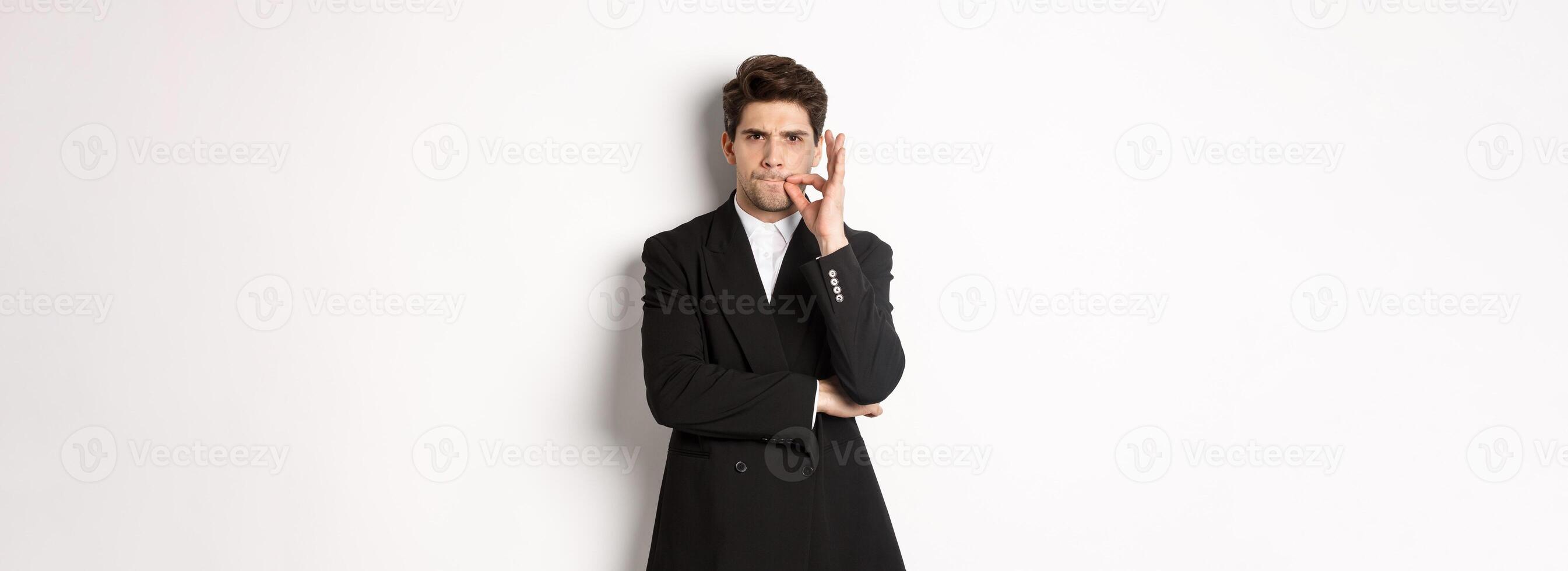 Image of serious businessman in black suit, zipping mouth and frowning, hiding secret, showing taboo sign, standing over white background photo