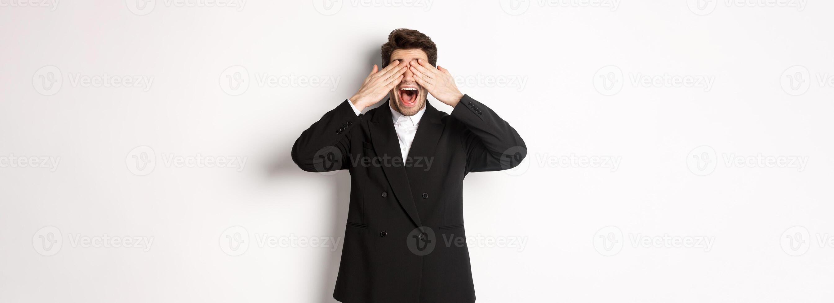 Image of handsome stylish man in black suit, waiting for christmas surprise, covering eyes with hands and smiling, anticipating presents, standing over white background photo