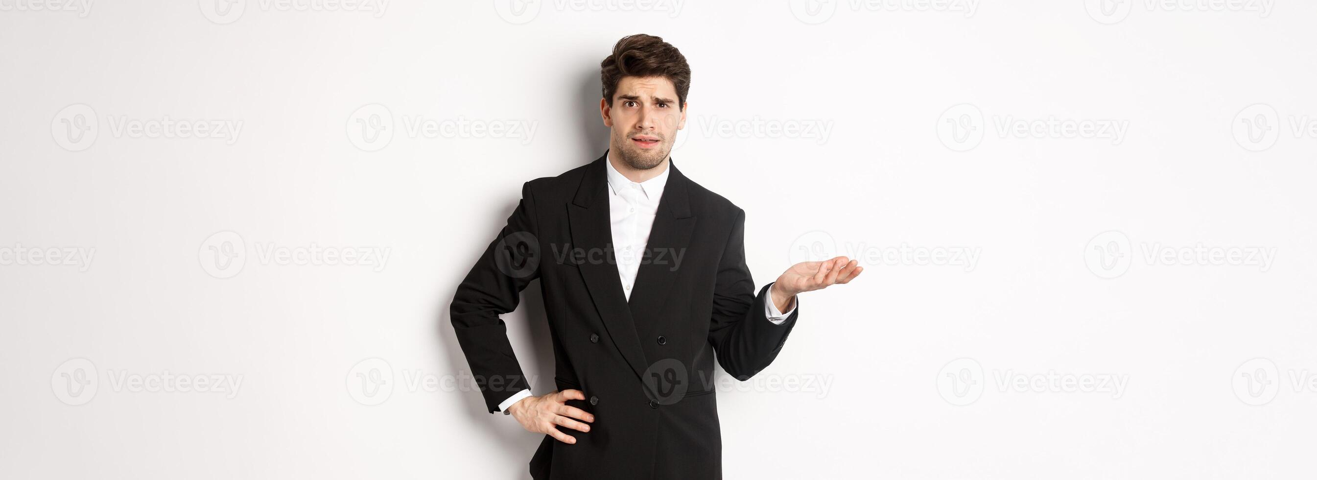 Image of confused handsome man in formal suit, raising hand and shrugging, cant understand something, standing against white background photo
