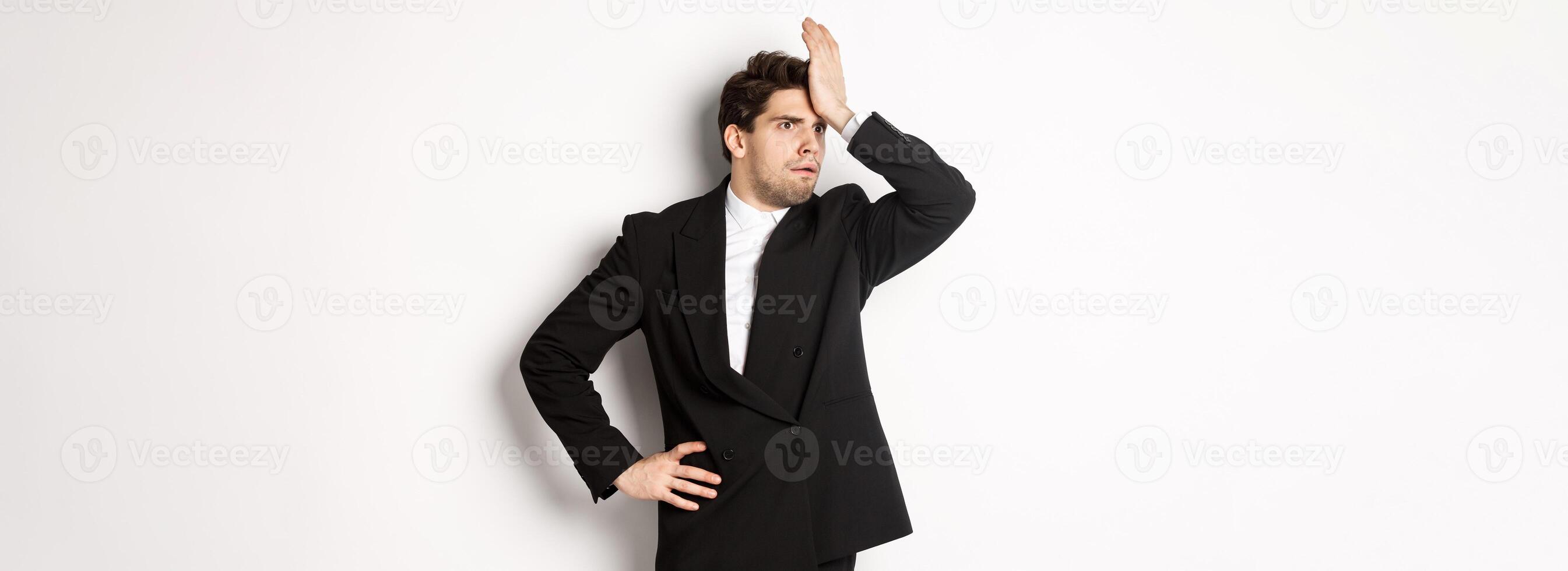 Portrait of shocked and distressed businessman in black suit, slap forehead and looking anxious, standing troubled over white background photo