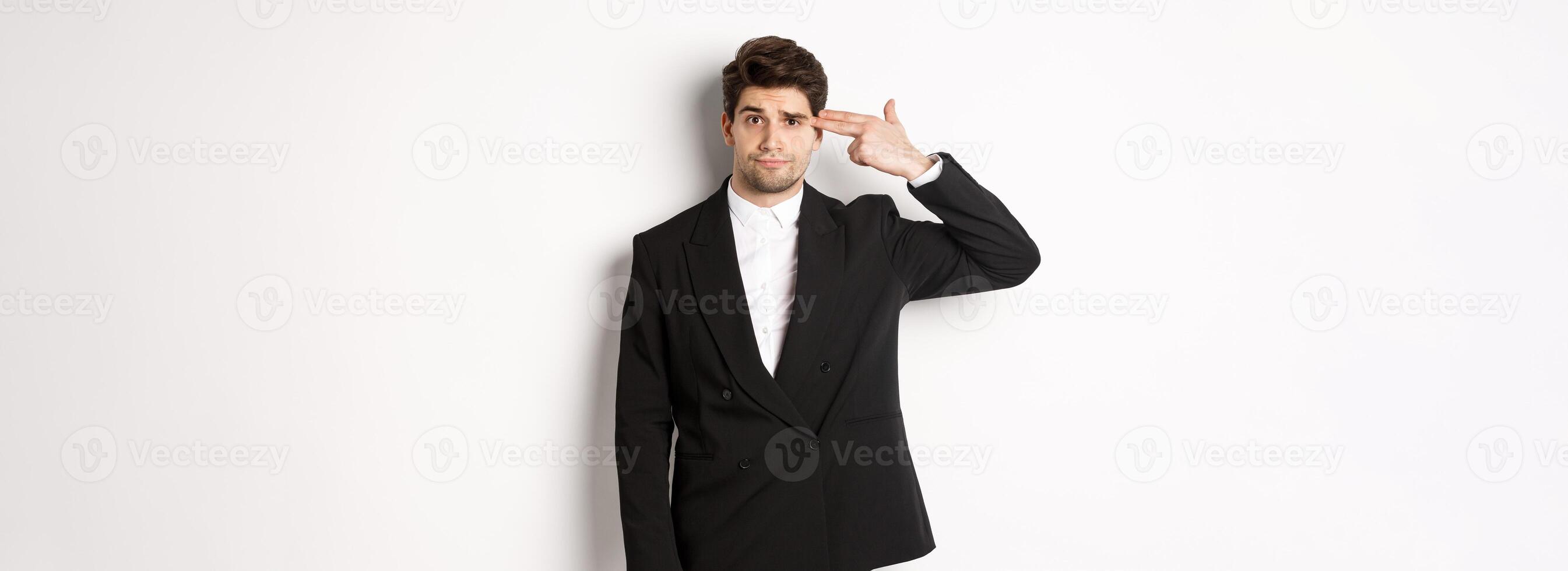 Portrait of distressed and troubled businessman in suit, pointing finger gun at head and looking bothered, shooting himself, standing against white background photo