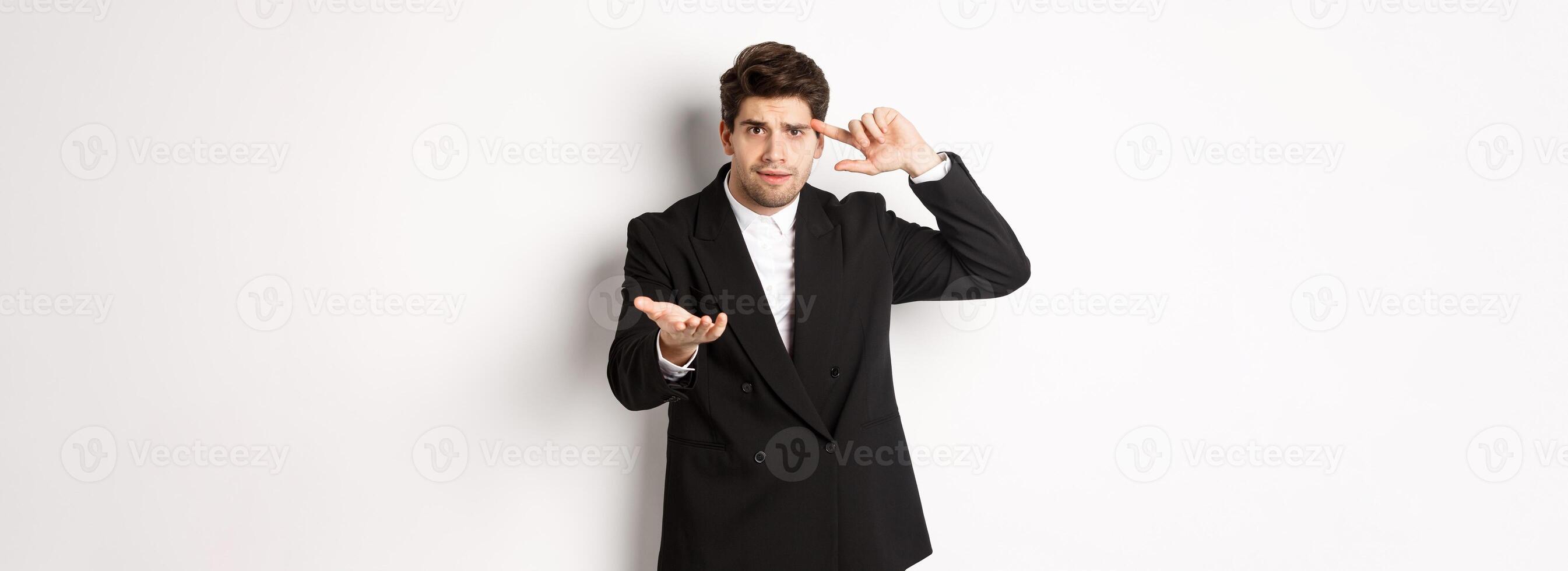Portrait of confused handsome businessman scolding employee, pointing at head and camera, standing disappointed over white background photo
