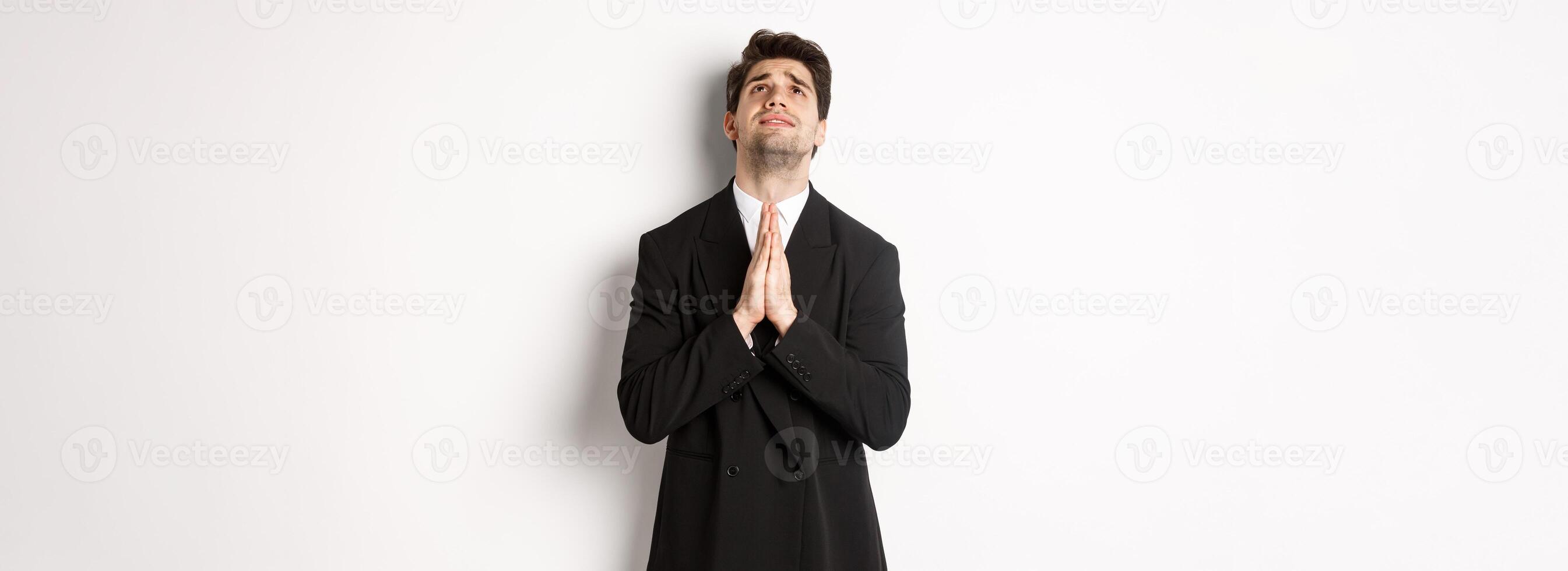 Troubled and hopeful man in black suit begging god, pleading and looking up, need help, standing over white background photo